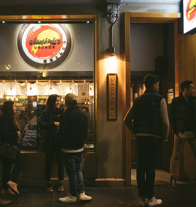 The Russell Street flagship at night, guests waiting beneath the シュジンコウ本店 sign